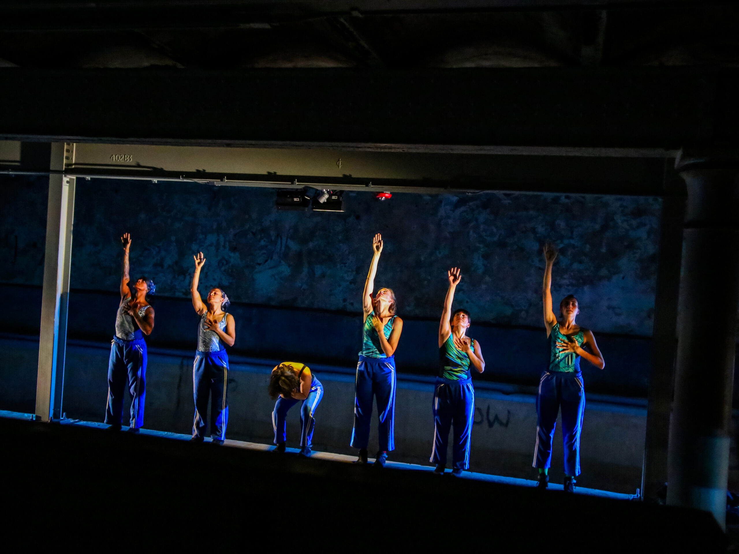 The six female dancers of The Naked Stark stand in a line facing upon a raised ledge above the subterranean Kelly Pool (not visible to the viewer). They face forward, wearing bright, electric green, patterned swimsuits and blue sweatpants. Five of the women gesture with their right arms stretched straight upward with their bent left arms resting on their chests. Each looks up toward her extended arm. The sixth woman forward bends over, her head and face into her torso, hiding. Precise side lighting illuminates their bodies with a silvery glow in the otherwise dark space.
