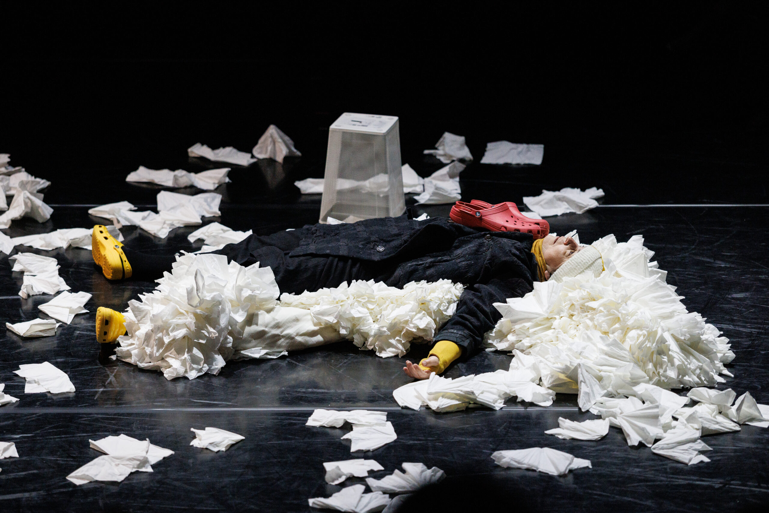 Tissues are scattered all over the black box theater floor. Upstage a white wire waste paper basket sits upside down and empty. On a huge pile of tissues lies a woman in black with yellow crocs, yellow sleeves sticking out of her coat, a yellow scarf and white and yellow pompom knit hat. She stares up at the ceiling. She has a pair of red crocs on her chest.
