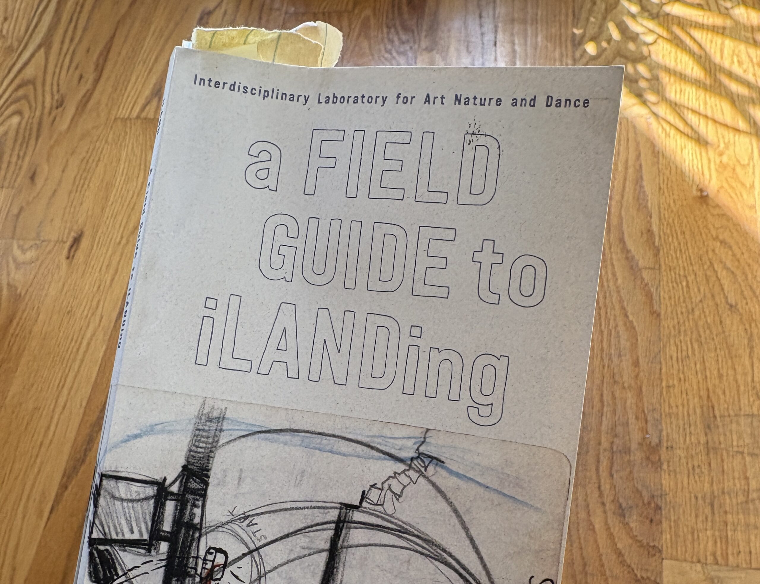 A hand holds a well-worn book over a sunlit wooden floor, with the shadow of a spider plant beside it. Torn yellow paper scraps stick out as bookmarks. The white cover reads “Interdisciplinary Laboratory of Art Nature and Dance, a Field Guide to iLanding,” above an abstract charcoal drawing of circles, arches, towers, scratches, and stacked squares.