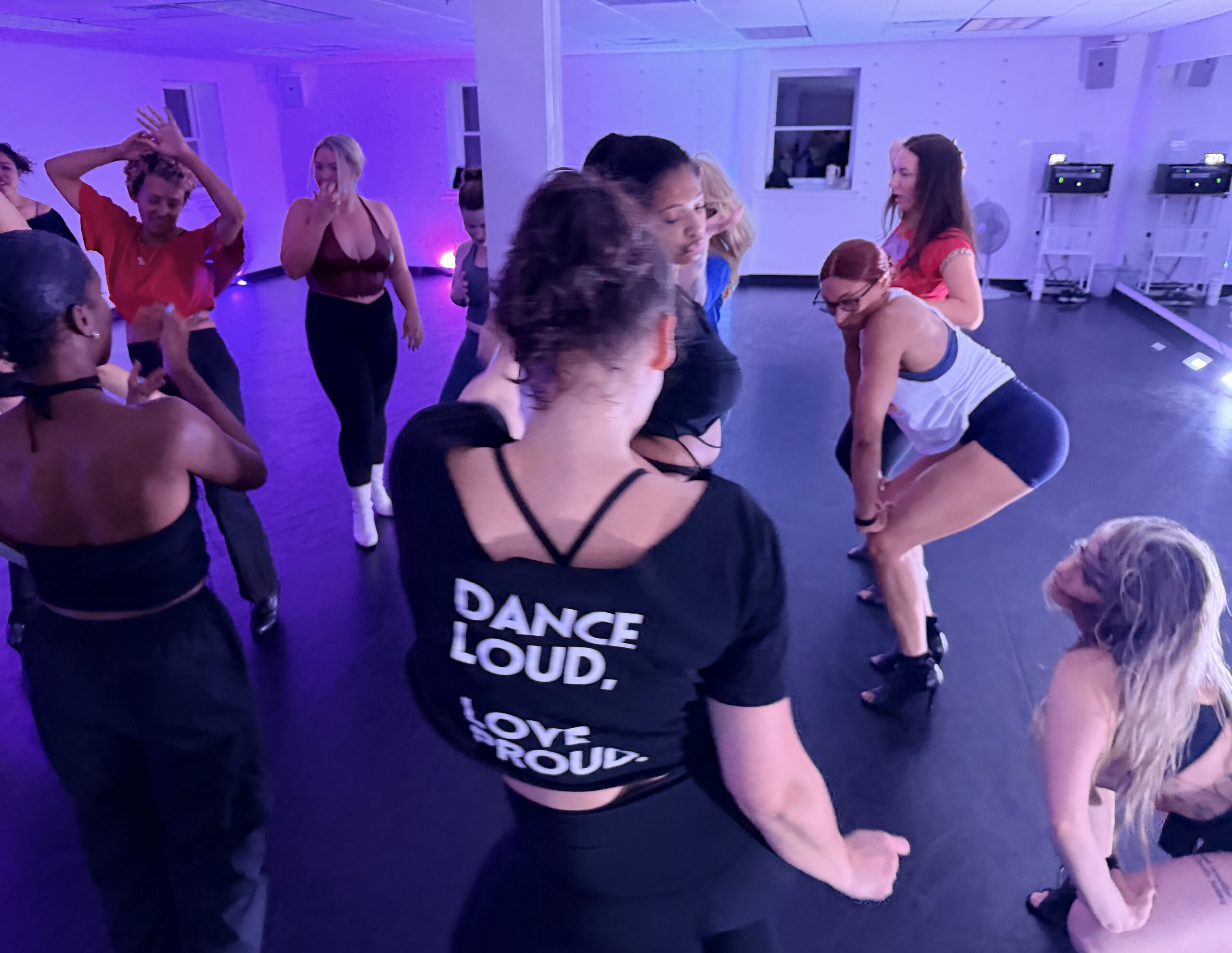 Overhead view of a dance class forming a circle under purple studio lights. A dancer in the center foreground wears a black t-shirt reading "DANCE LOUD, LOVE PROUD" while others around her cheer, dance, and watch.