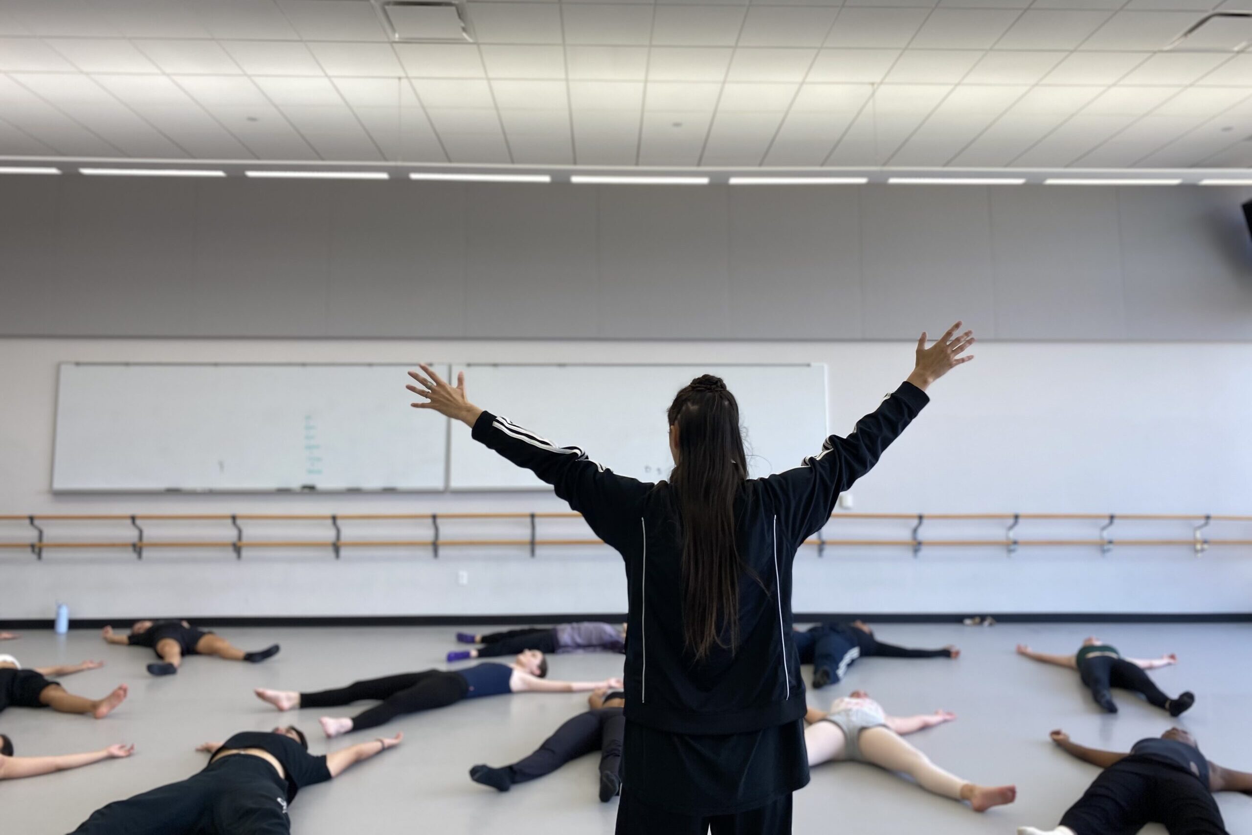 In a dance studio with white walls, a light gray marley floor, and two large whiteboards along the back wall, situated above a double-stacked wooden ballet barre, eleven college students lay supine with their arms raised and legs parted in an “X” shape on the ground. They lay at different angles, breathing deeply, following the instructions of the teacher at the front of the room. The instructor, tD writer Jennifer Passios, faces the students. She wears a black Adidas jacket. Her hair falls in a half-up-half down updo as her arms reach skyward in the same “X” gesture that the students make on the ground.