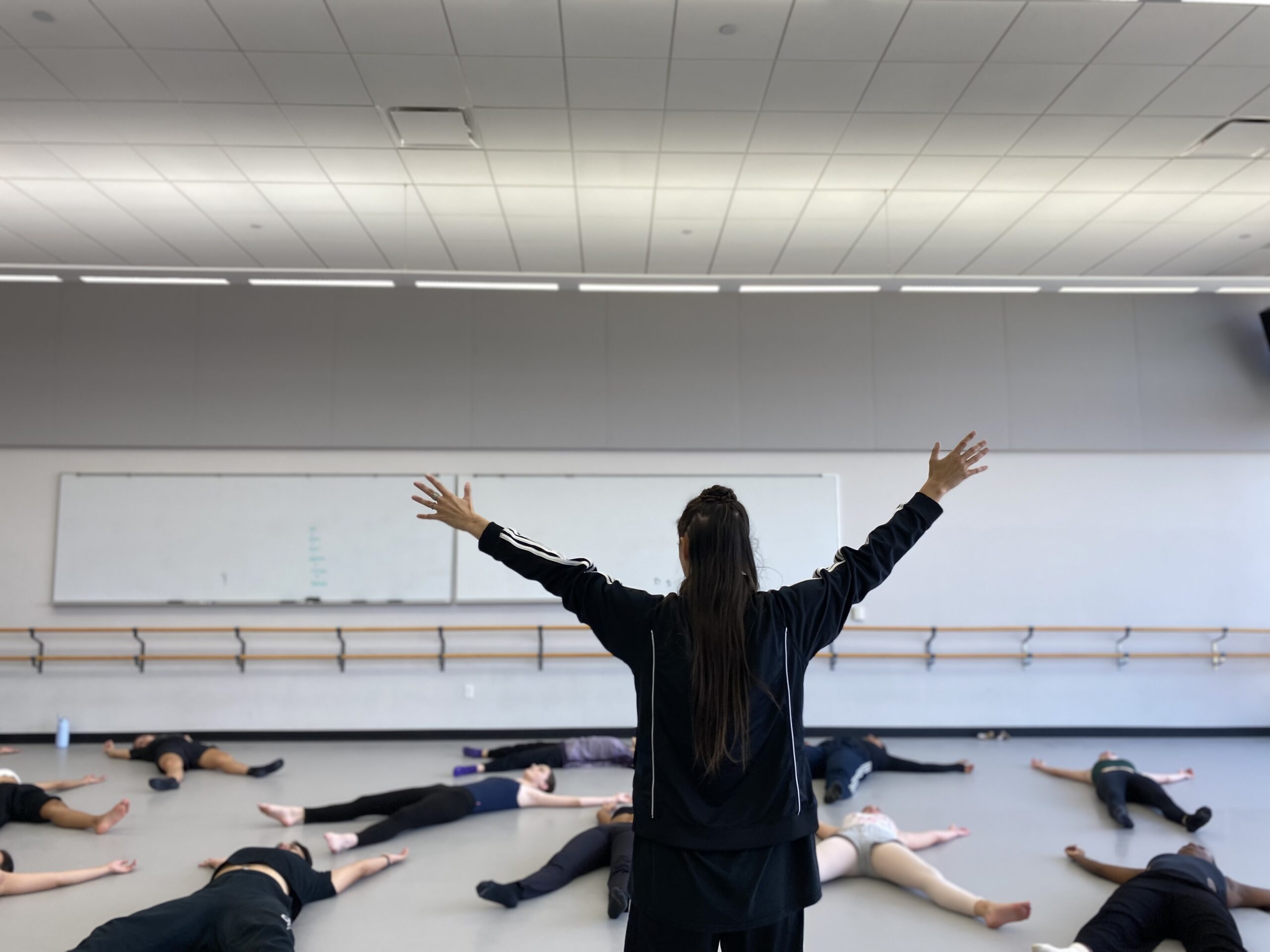 In a dance studio with white walls, a light gray marley floor, and two large whiteboards along the back wall, situated above a double-stacked wooden ballet barre, eleven college students lay supine with their arms raised and legs parted in an “X” shape on the ground. They lay at different angles, breathing deeply, following the instructions of the teacher at the front of the room. The instructor, tD writer Jennifer Passios, faces the students. She wears a black Adidas jacket. Her hair falls in a half-up-half down updo as her arms reach skyward in the same “X” gesture that the students make on the ground.