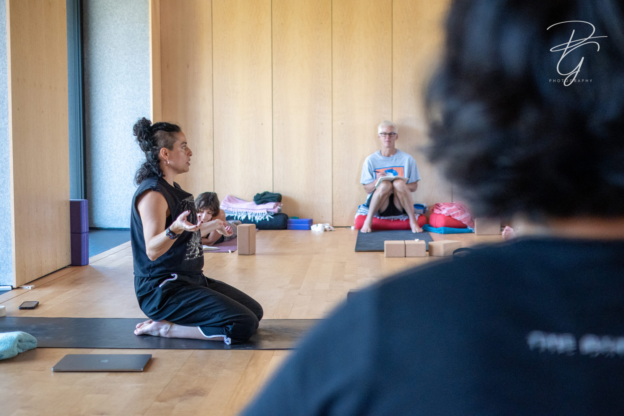 Zaquia Salinas kneels on a yoga mat in a naturally-lit studio with wood walls and flooring. She embodies a grounded, punk energy, with a half-shaved head and an all-black outfit of a muscle shirt and capri-length workout pants. Her hands gesture out from her body as she speaks to a class of participants who are similarly seated on yoga mats with blocks surrounding them. The participants include one black-haired individual whose head is foregrounded in the image, one white-haired individual in t-shirt and shorts on the opposite side of the room, and a brown-haired person laying down on their stomach reading from their journal as Zaquia speaks.