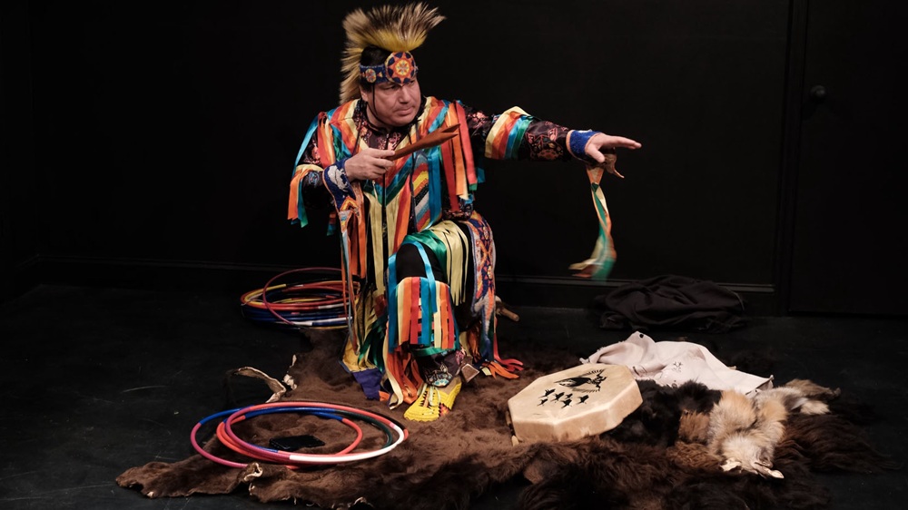 Delwin Elk Bear Fiddler kneels with one arm outstretched to the left and the other bent across his body holding a wooden pipe. Fiddler wears bright attire with multicolored strips of ribbons and a yellow feathered headdress. He kneels on a brown animal hide near a drum and several piles of plastic hoops in a blackbox theater.