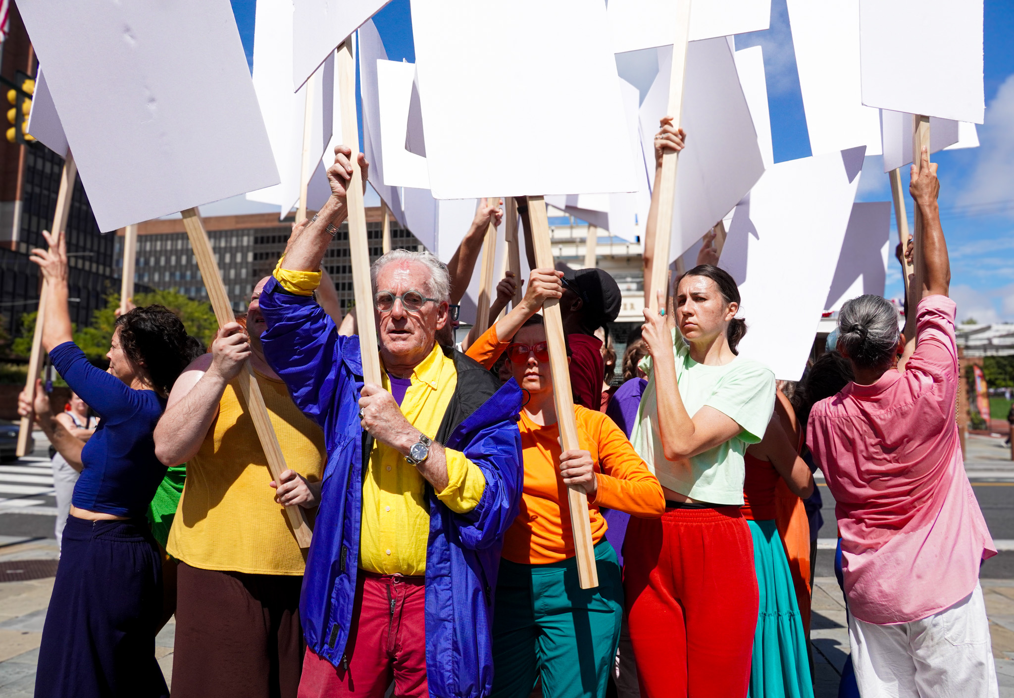 Dancers stand close together, forming a clump. They face in all directions, their bodies back to back, front to back, and back to side. Each one grips a wooden post holding high attached white posters. They wear bright yellows, purples, reds, blues, pinks, teals, and oranges. Some are visibly sweaty. Dancers facing the camera look serious and a bit fatigued. Glimpses of blue sky, clouds, a street, and city buildings fill the cracks between their blank signs.