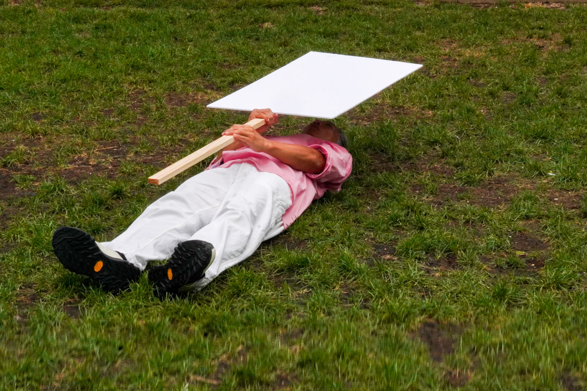 A man in bladk shoes, white pants, and a pink button up shirt lies on the ground on green grass. He holds a blank sign on a wooden pole horizontally over his body