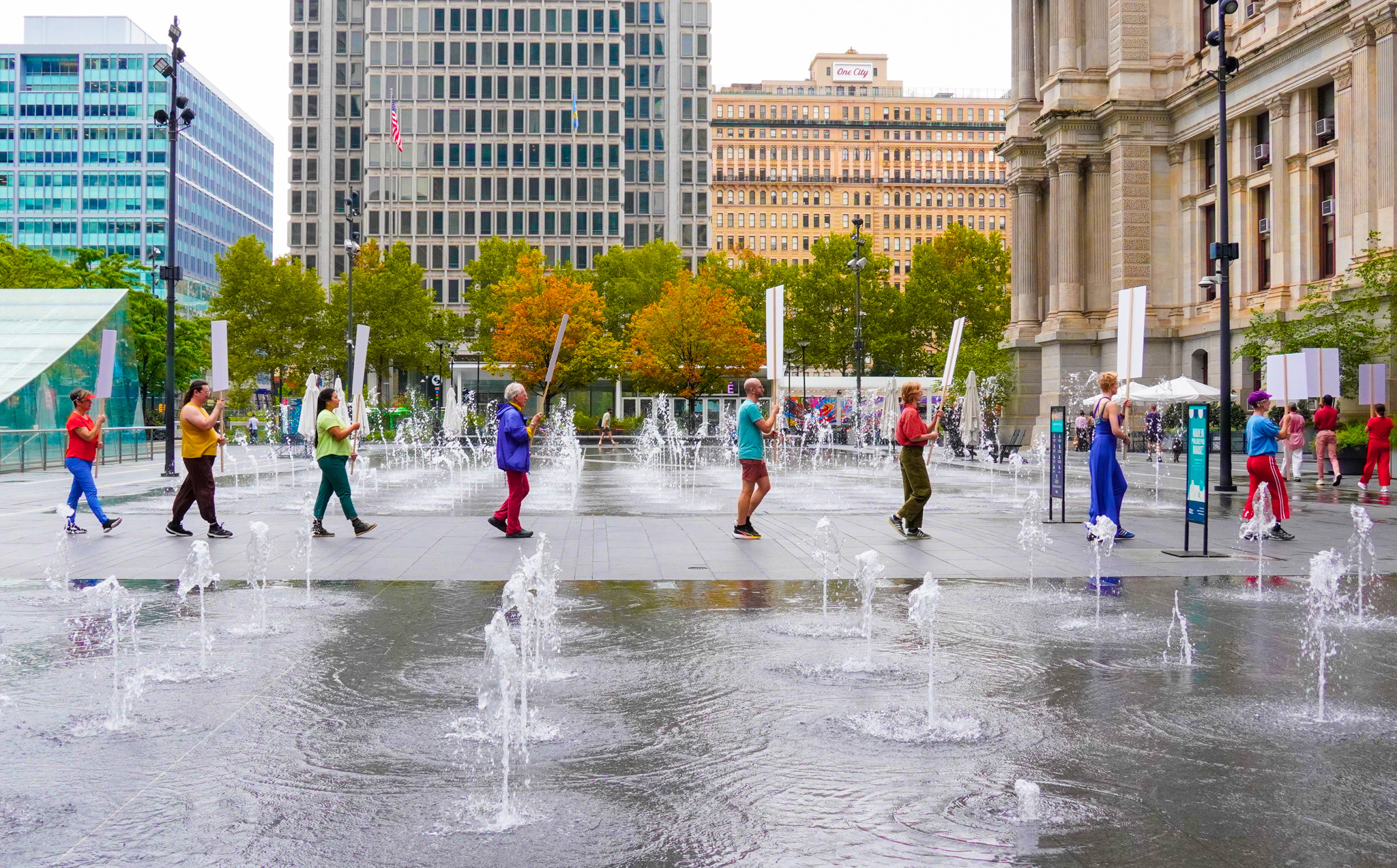A line of people in bright, solid colored clothes walk across Dilworth Park. From this camera angle, we catch the sides of blank white signs they hold up, and see three to eight feet of space between each marcher. They pass a grid of sprouting water fountains in the foreground and the background. Behind them, large downtown buildings, both modern metal and glass, and old stone, loom.