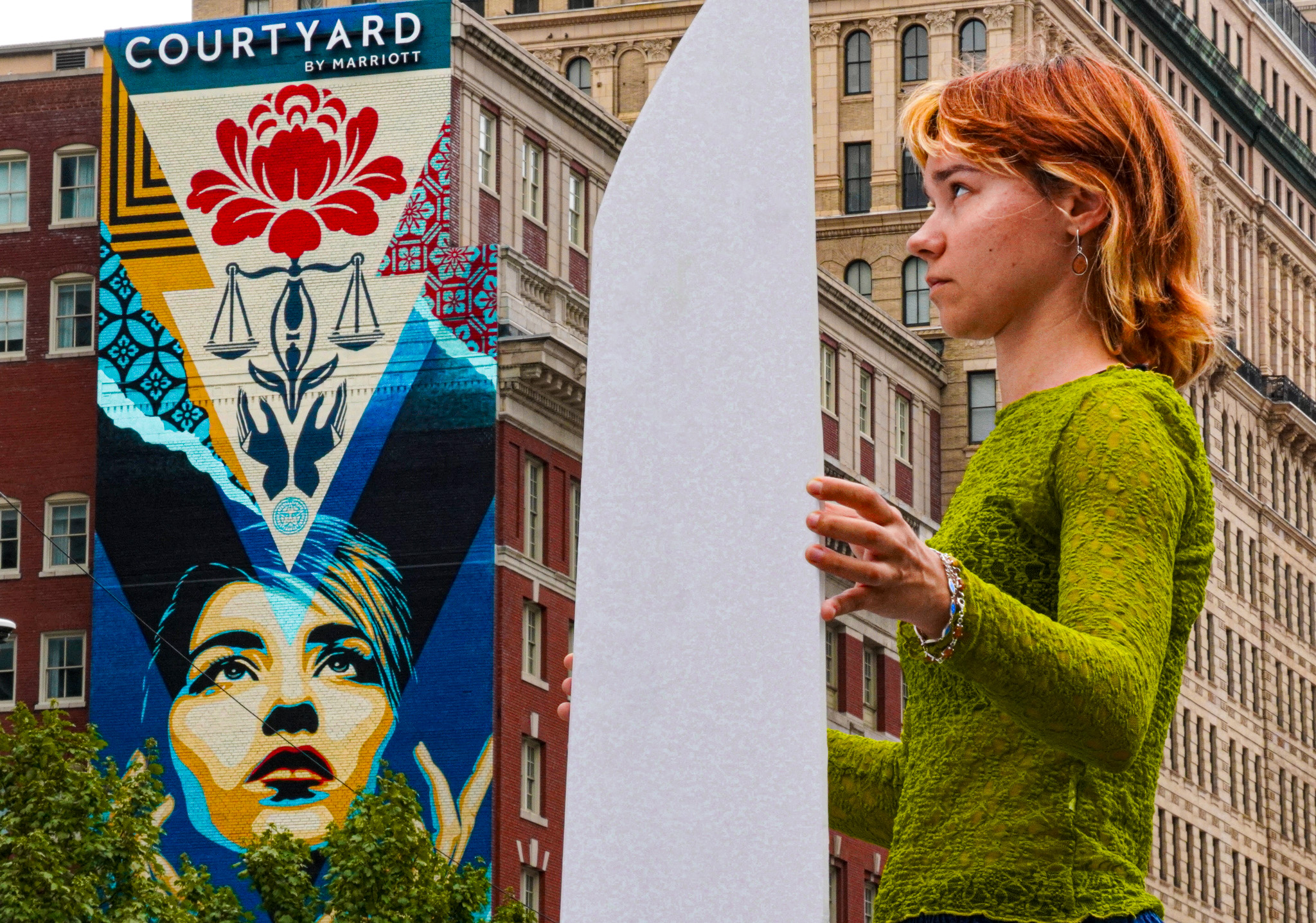 A young person with shoudler length red hair stands in a green long sleeve shirt hodling a blank white sign. onthe left a large mural of a female face looking up, and scales and lotus flower.