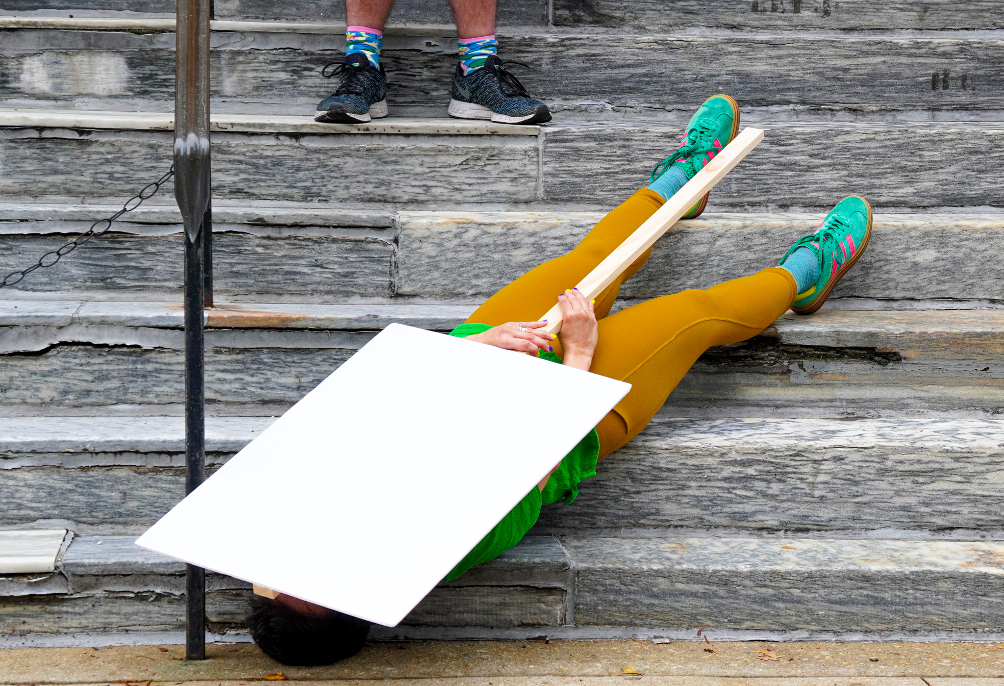 a person wearing mustard yellow pants and bright blue green shows lies on their back on a flight of stone stairs, their head pointing down. They hold a blank sign over their body.