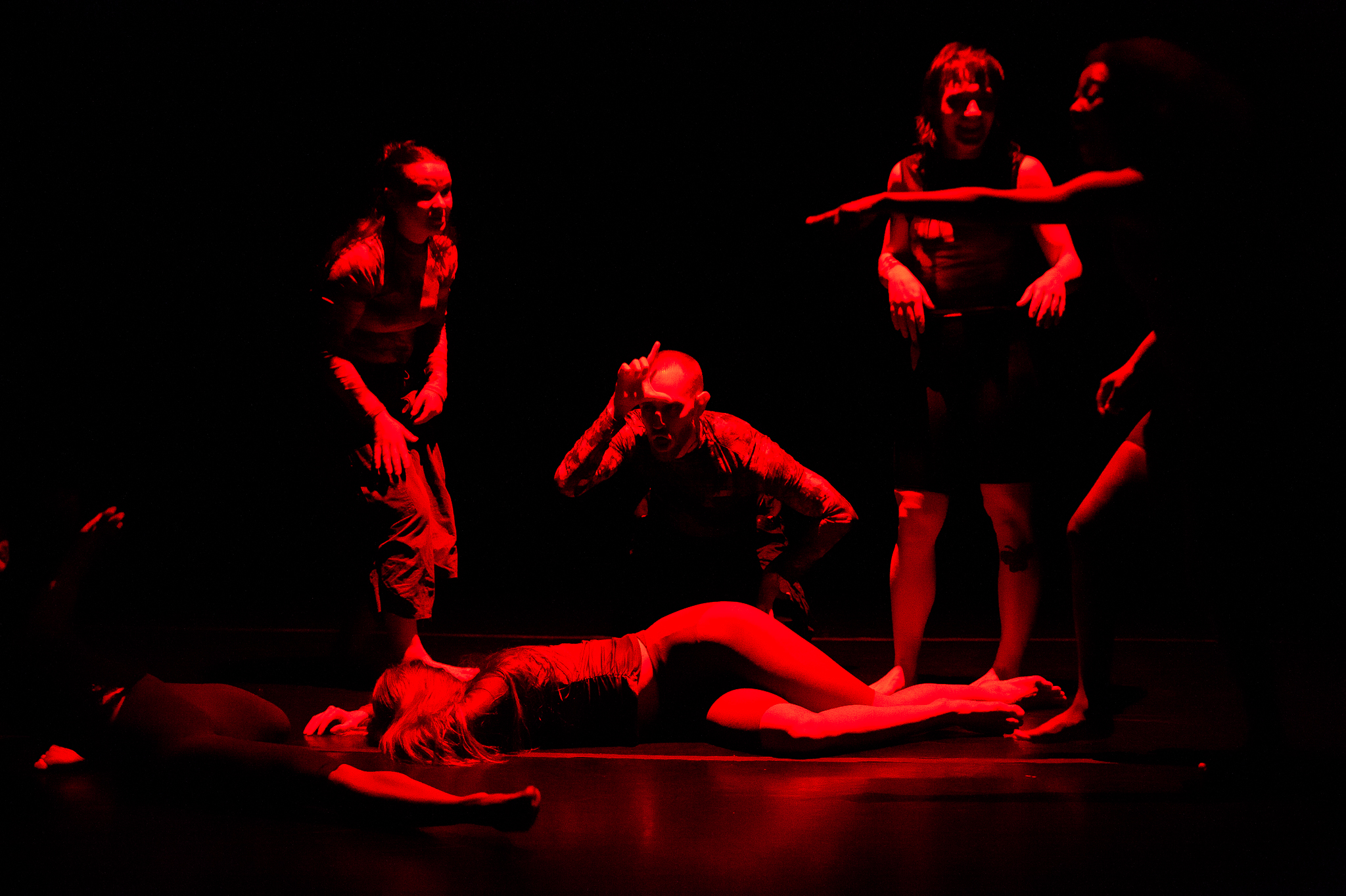 The light’s red, the stage black. Dramatic. Devilish. Contrast and shadow are high on the six people in frame. Dead center, a dancer lays. Around her, four people point, laugh, and crouch over her body making the “loser” L on their forehead. In the bottom left corner, someone's leg, hip, and reaching hand are just in frame.