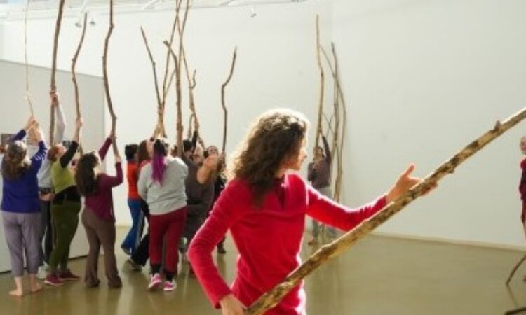 Dancers rehearse for the closing performance of Merián Soto’s exhibition last January celebrating her 25 years at Temple University. Marion Ramírez is center. Soto is to the right, overseeing the rehearsal and also participating. Each dancer is dressed in colorful, casual exercise clothing and holds a long branch vertically along their body, arms raised above their head and branch extended into the sky. From her vantage point, Soto is slightly removed; she is smiling.