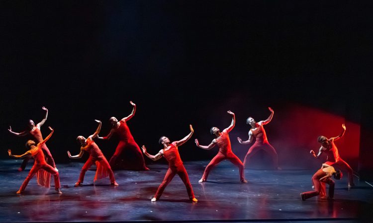 9 dancers dressed in red spread across the stage. 8 of these dancers’ arms part above their heads, eyes tilting up as if to look towards the heavens hoping for strength. One dancer is an exception, keeping their head down to the ground while in a shallow diagonal lunge.