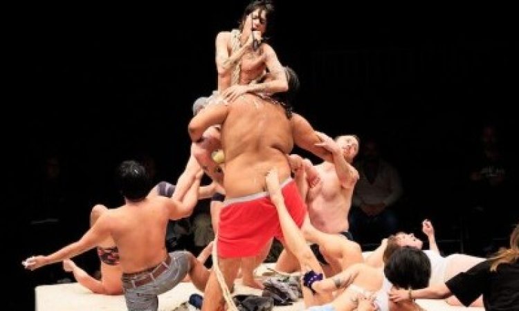 A mass of performers writhe on a square white platform. Some of the performers kneel while others lay or drape themselves atop one another. One shirtless performer rises above the others, held by a Black performer wearing red boxer shorts. The performer held aloft croons into a microphone; a rope is draped over the shoulder of the arm holding the mic.