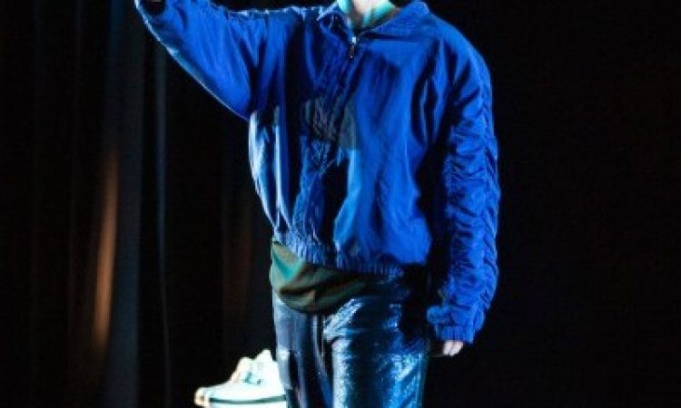 white person wears an astronaut helmet with the face shield down, though it doesn’t quite cover their chin. They stand with one foot in front of the other and hold up an arm, softly opening their palm to face forward as if waving or saying “hold up”. Turquoise light makes shapes on their open palm and their neck. They’re dressed in textures of blue, glimmering pants and a blue bomber with cinched ruffled seams along the arms. Two stage blocks rest behind them, one with a pair of plush white boots set atop.