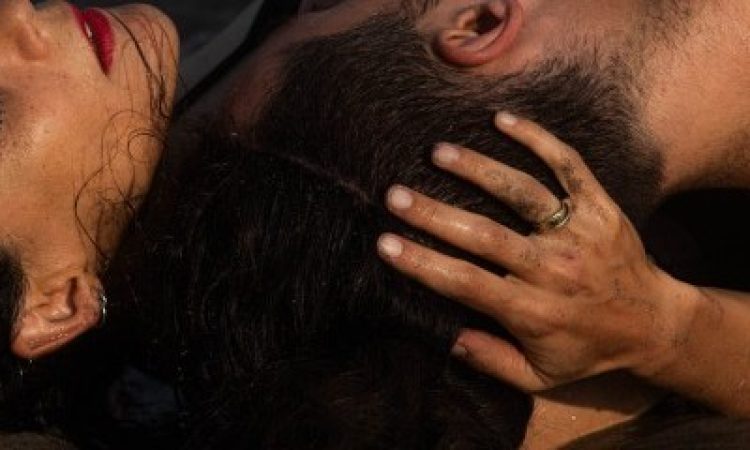 "hug" - Mor Mendel, a woman with black hair, hugs the head of David Guzman close to her shoulder, as the two lie on the wet sand. On her hand we see a gold ring.