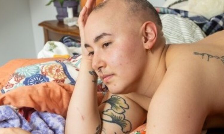 Ada Fujita lays shirtless on a bed unmade with orange and multicolored blankets. They rest their head on their hand and gaze softly down and to the side. They take up most of the image frame, visible from the waist up, their buzzed head center frame. A polished wood bedside table with potted plants, more blankets, and a stuffed animal are blurry behind them. Ada’s tattoos of leaves and line drawings interplay with the patterns of their bedding.