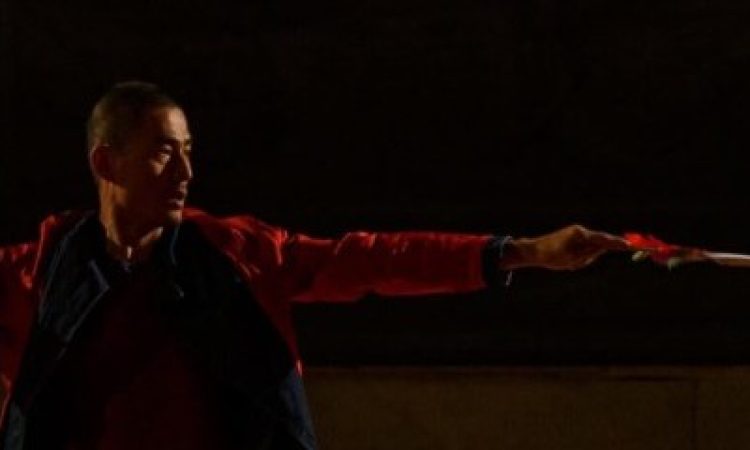 An artist stretches their arms side to side with their body positioned towards the camera. They are looking in the direction of one extended arm and appear to be holding a wand or blade in their hand.