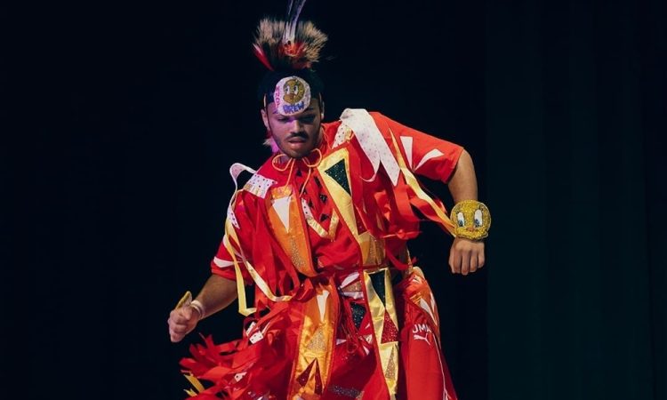 A dancer wears a feathered head piece and bright red garment with yellow, orange, black, and white strips of fabrics that dangle as they dance in front of a black backdrop.