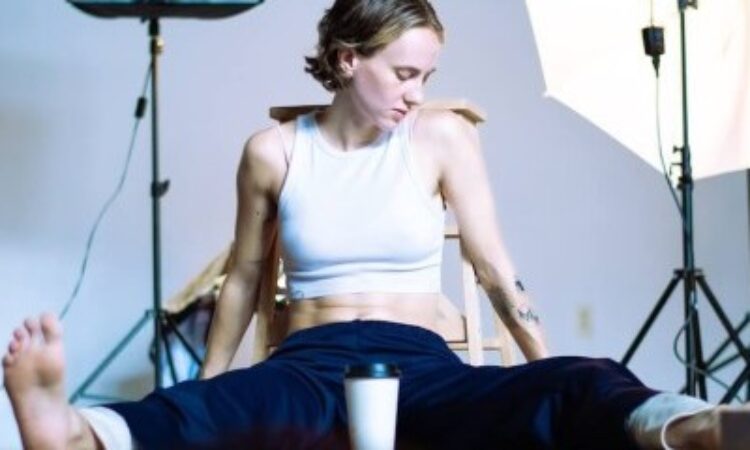 The performer, marisa illingworth, sits on the floor in a straddle with their body facing the camera. They are wearing a white crop top and navy sweat pants and have bare feet. A coffee cup sits in between their legs, white with a black lid, anonymous.Tattoos are visible on their arms. They look down and away from the camera. Behind them is photography lighting equipment; one of the lights is lit, putting the performer in the spotlight.