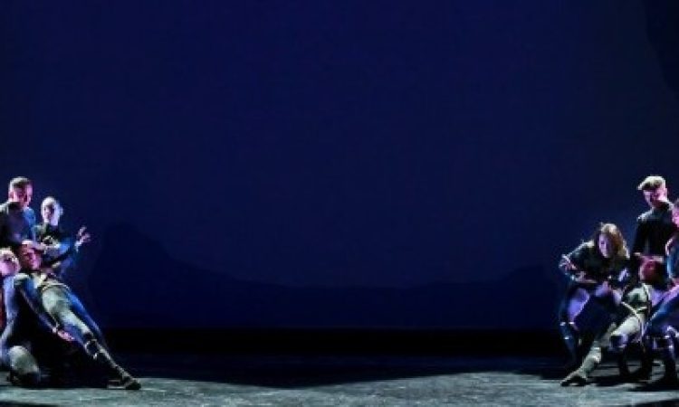 two groups of dancers split the stage, each one catching a falling dancers body. they all wear black with gold accents, the stage is lit in two large diagonal, crossing rectangles.