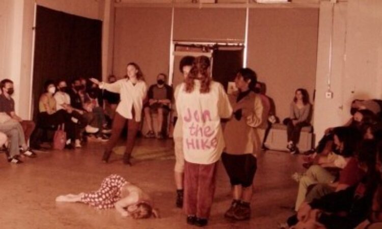 A warm, slightly grainy, tinted photo of an in-the-round white-walled performance space. At the center of the room a performer in polka-dot pants lies on the ground. Above her, three performers gather, one wearing a shirt that says “Join the Hike.” To their left, Corinne Jones – wearing a white button-up and brown tights – gestures out to the audience with an open arm.