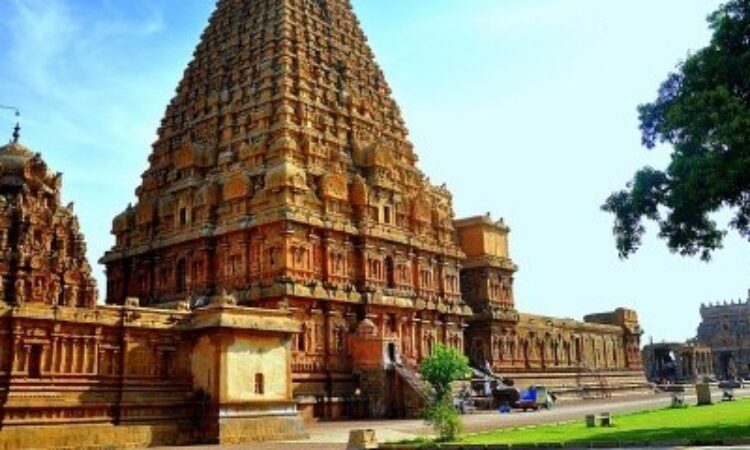 A view of the outside of the Brihadeeswara temple shows two large golden turrets with many intricate carvings, surrounded by a similarly carved wall. There is a bright blue sky behind the temple and a large tree on the other side of the image. 