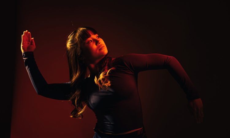 Jazlyn Tan, clad in a long-sleeve black top, leans her weight to one side. Her hands are blades; one arm in at a ninety-degree angle up, the other an inversion. Her gaze is focused and serious.