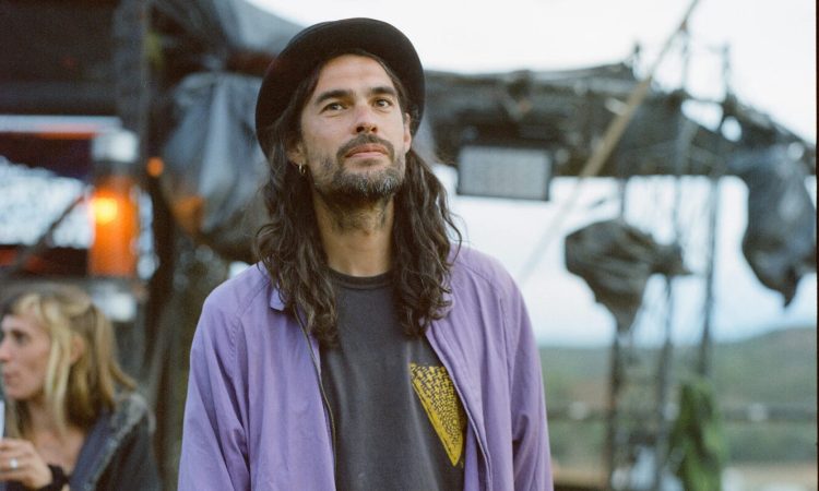 A young man stands relaxed in front of what looks like some kind of set in the middle of the desert, a woman (out of focus) with blonde hair stands behind him to his right. The man has long black hair that trails down to his collarbones, and matches the color of his beard. His hat is tilted towards the back of his head, and he wears a light blue jacket over a black tee-shirt, and leopard-print pants.