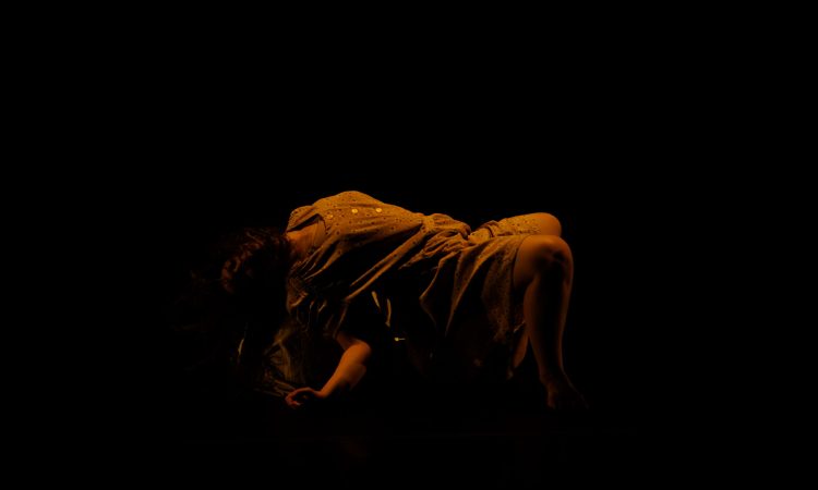 In a sparsely lit space, a dancer with long dark hair lifts her hips up from the ground in a long cotton dress. Her hair covers her face, and she pushes up from the ground with her right forearm.