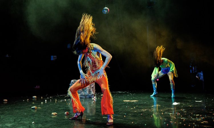 On a dark smokey stage littered with fruit loops and rocks, two dancers whip their hair forward from a wide second position plie (legs wide, turned out, and bent), their hands smacking their thighs. The dancers are dressed like it’s the 70s and they’re going to the club: one in an orange halter jumpsuit with sparkly fringe, the other wearing tall silver heeled boots, purple booty shorts, and a fluorescent green shirt with flared sleeves.