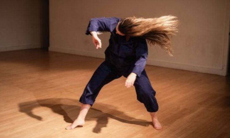 Kay MacMaster wearing blue coveralls stands in the center of a wood paneled performance space. Their body is folded forward, knees slightly bent, and the head tossed sideways with their long blonde hair dramatically fanning out, capturing a feeling of wild abandon and emotional release.