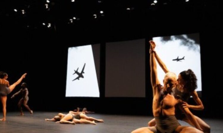 Three groups of dancers appear onstage in front of projected images of planes. To the left, a woman extends her right leg high while a man nearby bends his knees and has his arms out from his waist. On the floor, in the middle, three figures lie on their right sides. Closest to us and on the right, a man squats deeply with his arms extended diagonally while a woman bends her knees behind him, holding his wrists high.