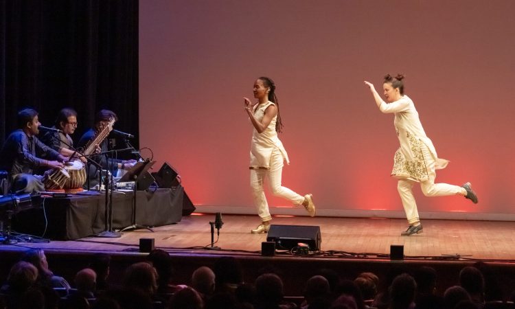 From left to right, dancers Dormeshia, Rachna Nivas, Rukhmani Mehta and Michelle Dorrance. They are in motion. Dormeshia and Dorrance wear white pants, thigh length white tunics, and tap shoes. Nivas and Mehta wear white leggings, long white dresses with golden details on the skirts and bodices. They have bands of bells around their ankles and are barefoot. The tap dancers have a quality of bending and sending energy into the floor. The Kathak dancers are lifted, arms raised, poised.