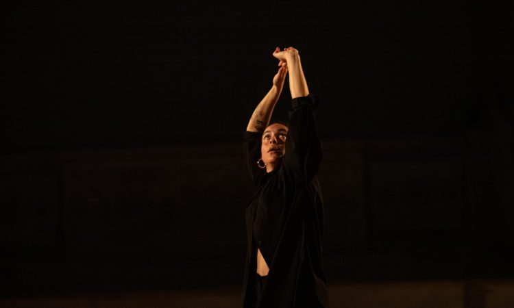 A performance image of Zaquia Salinas, taken during her piece, A Call to Prayer. She wears large hoop earrings and an all-black outfit, with a shirt that wraps and opens around her stomach. Her gaze is directed upwards with a focused expression that manages to feel both wondrous and apprehensive. Her arms snake above her head asymmetrically, almost as if her hands are pushing the sky away.