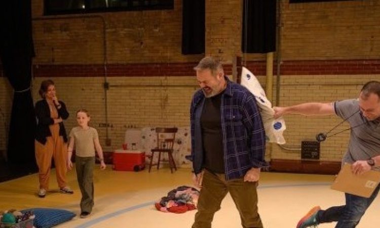Alex Torra winces as Benjamin Camp hits him on the back with a patterned pillow. Benjamin is jumping with the force of his throw, clutching a clipboard and wearing a stopwatch around his neck. Sydney Camp, Benjamin’s daughter, calmly walks between a basket of toys and a pile of clothes behind them. Rachel Camp, Benjamin’s sister, watches behind Sydney, a hand in front of her mouth hiding an amused smile.