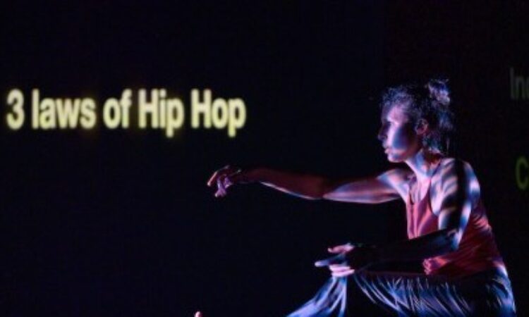 One dancer balances her bent right leg, left leg lifted, out stretched to match her opposite arm.Words to the left of the frame written on the back wall read “3 laws of hip hop.” The rest of the stage is nearly black.