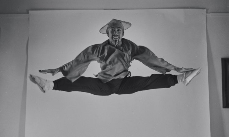 Black-and-white photo from 1954 of George Lee, a Chinese man, performing a mid-air jump split. He wears theatrical costume and a painted beard, with exaggerated facial features in yellowface makeup.