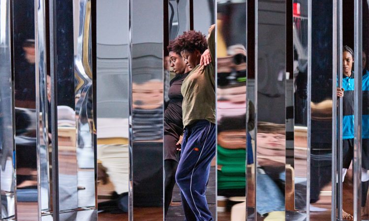 Performers Kris Lee and AJ Wilmore, both Black women, stand inside a metal structure with mirrored panels. Lee wears a black shirt and pants. Her left arm is draped over Wilmore's shoulder, and she looks straight ahead. Wilmore wears an olive-green shirt and navy-blue sweatpants. Her left knee is bent, and her left arm is raised, and she looks down. Bria Bacon, in a blue-and-black striped t-shirt, black leggings, and white knee pads, is in the background, her hand on the structure.