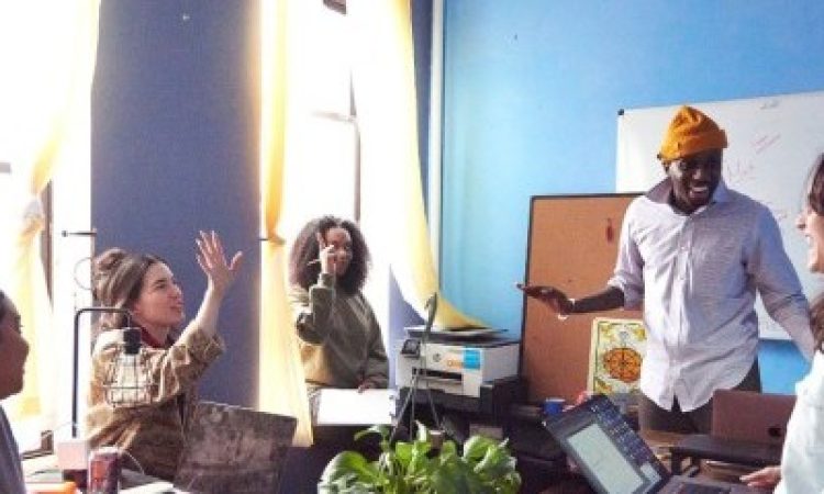 Raja Feather Kelly and the feath3r theory team gather with open laptops and poised notebooks in a sun soaked room with blue walls. Raja stands with his right arm bent at the elbow, smiling with teeth parted as if beginning to laugh mid idea. Two of the other team members have their hands raised, ready to contribute to the conversation. The tools of brainstorming – a whiteboard, a corkboard, a hanging plant that isn’t hanging, pens, books, and a conveniently placed LaCroix – furnish the meeting space.