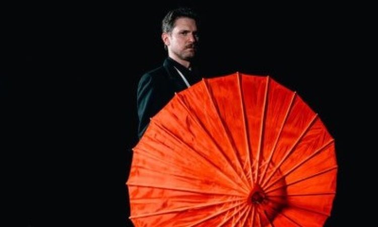 Against a black background, Schreiben wearing a black suit stands partially obscured by the vibrant red parasol he is holding.