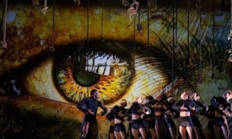 he image features glowing amber eyes projected onto the backdrop of the stage. On the sun-dappled-like stage, a group of BalletX dancers stands tight together, mirroring the character Jack’s command to lift their strong and angular arms to their hearts. Dressed in black, slim-cropped jackets and shorts, the dancers’ shrugged shoulders convey a sense of suspense or reverence. The atmospheric lighting, combined with the haunting gaze of the eyes, creates a mood of mystery and psychological intensity, reinforcing the ballet’s themes of surveillance, power, and primal instinct.