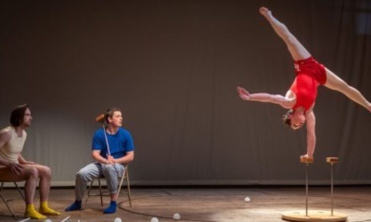 Liam Bradley and Kevin Flanagan watch Sierra Rhoades Nicholls intently from their position to the side of the stage, seated in folding chairs. Nicholls supports herself upside-down with one hand gripping a block that is balanced atop a silver rod. Her other arm is extended parallel to the floor at her side and her legs are extended in a straddle above her head.