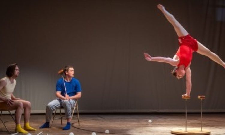 Liam Bradley and Kevin Flanagan watch Sierra Rhoades Nicholls intently from their position to the side of the stage, seated in folding chairs. Nicholls supports herself upside-down with one hand gripping a block that is balanced atop a silver rod. Her other arm is extended parallel to the floor at her side and her legs are extended in a straddle above her head.