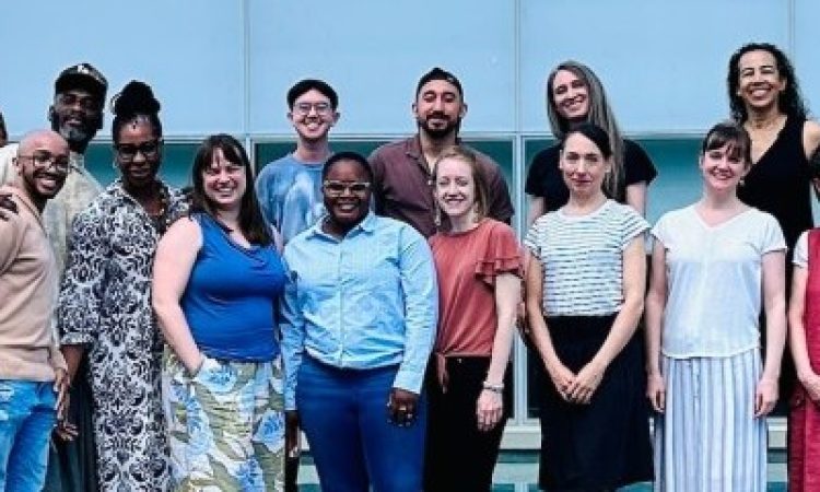 A group of dance writers stands in two rows facing the camera. They are wearing bright colors, smiling, and squinting into the sunlight.