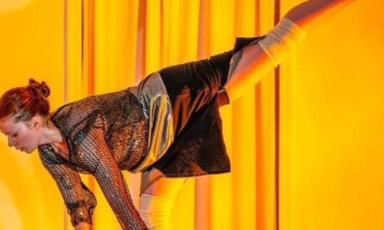 Swathed in warm yellow light, Amanda Rattigan stands on one leg, hinging deeply at her hips. She makes one long diagonal from her toes, that reach high behind her, to her eyes, that focus towards the ground. She extends her arms toward her standing foot, palms turned forward, fingers soft. She wears a sparkling, netted black long sleeve over a shimmering silver tank top with a black skirt that falls above her knees, her knee pads visible.