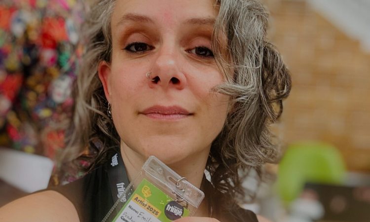 Emilee Lord, in a black tank top, smiles into the camera, her grey and brown curly hair framing her face. She holds the participant badge for Edinburgh Fringe in front of her chin.