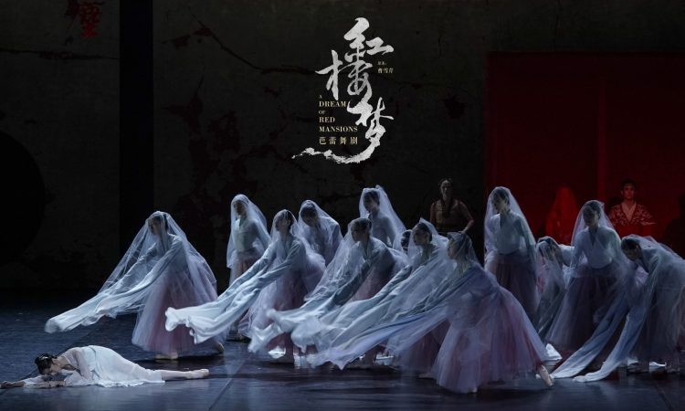 This is a haunting stage moment. Downstage left, Lin Daiyu lies collapsed on the floor, her pale costume blending into the cool blue-gray lighting, evoking fragility and lifelessness. Surrounding her, a semicircle of female dancers in white veils and soft pink skirts lean forward in unison, their long, flowing sleeves extended toward her like drifting currents or mourning spirits. The translucent veils soften their faces, creating an ethereal, ghostlike presence reminiscent of the Wilis from Giselle. The overall composition—low, reaching arms, rippling fabric, and a grounded central figure—creates a visual image of collective grief, where movement, costume, and spatial design merge into a poetic expression of mourning.