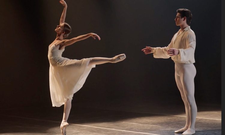 Oksana Maslova balances on pointe in first arabesque, leaning back toward Sterling Baca, who stands behind her in support. She faces upward, arms extended, with a poised, expressive posture; both dancers’ faces convey stoic grief.