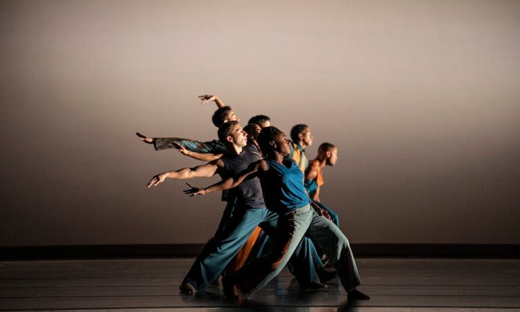 Alvin Ailey American Dance Theater dancers stand in a vertical line on stage. The dancer at the front of the line, furthest downstage, obscures the others behind her. The dancers wear a mix of blue and grey costumes. They lean back in a split leg stance, extending an arm behind themselves.