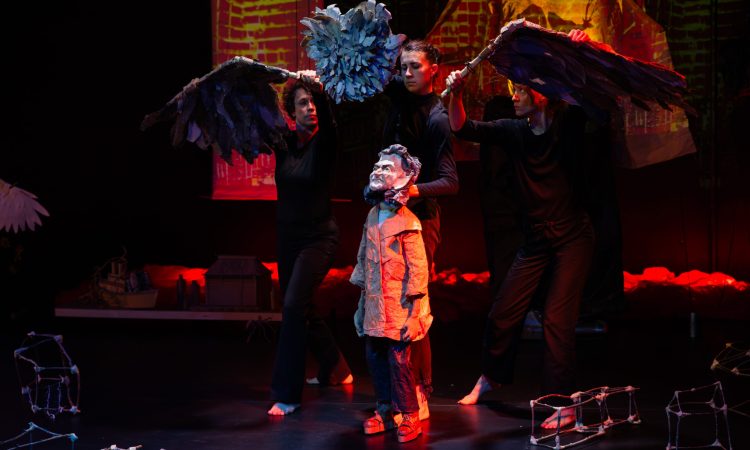 A papier-mache puppet of Kea Tawana is overshadowed by two massive disconnected owl wings and an eyeless owl head soaring above her, held by the puppetering cast dressed in all black. The Kea puppet is wearing an oversized trench coat, trousers and sandals. Parts of her ark lie scattered around her, made of twigs and tape. There is a spotlight over her, and the rest of the stage is darkened.